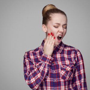 Tooth pain. unhappy blonde girl in red, pink checkered shirt, collected bun hairstyle, makeup standing touching her painful tooth with open mouth. indoor studio shot. isolated on gray background
