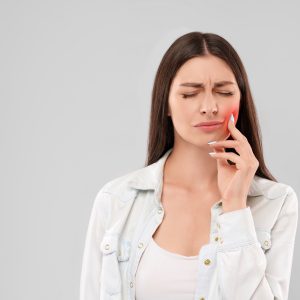 Young caucasian woman in white shirt over gray isolated background touching mouth with hand with painful expression because of toothache. Front view of brunette with closed eyes. Dentist concept.